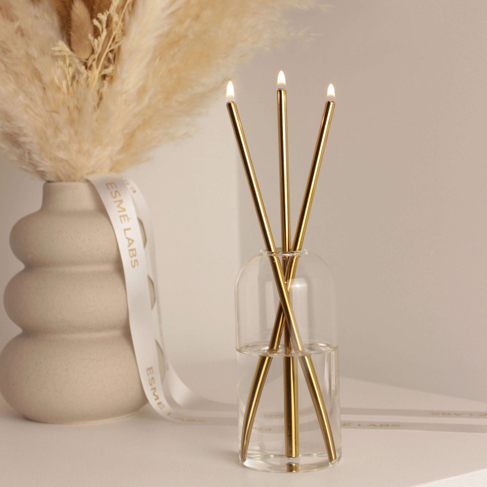 Long-lasting Esmé Labs candle in a clear glass vase with three lit gold wicks, next to a beige ceramic vase with dried pampas. Neutral background.