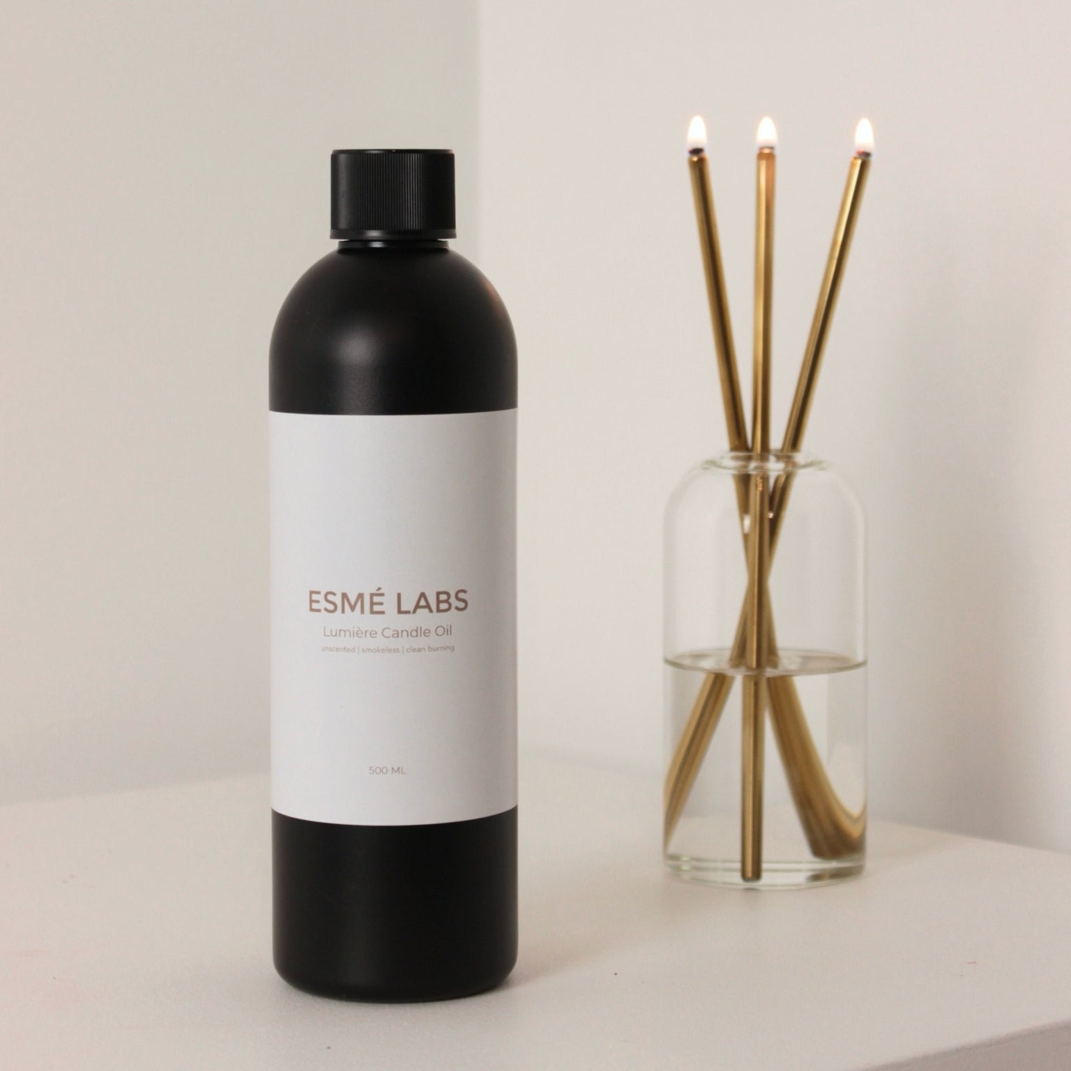 Black bottle with white label labeled 'ESME LABS' Lumiere Candle Oil next to a Lumiere Candle glass vase with gold wicks on a light background